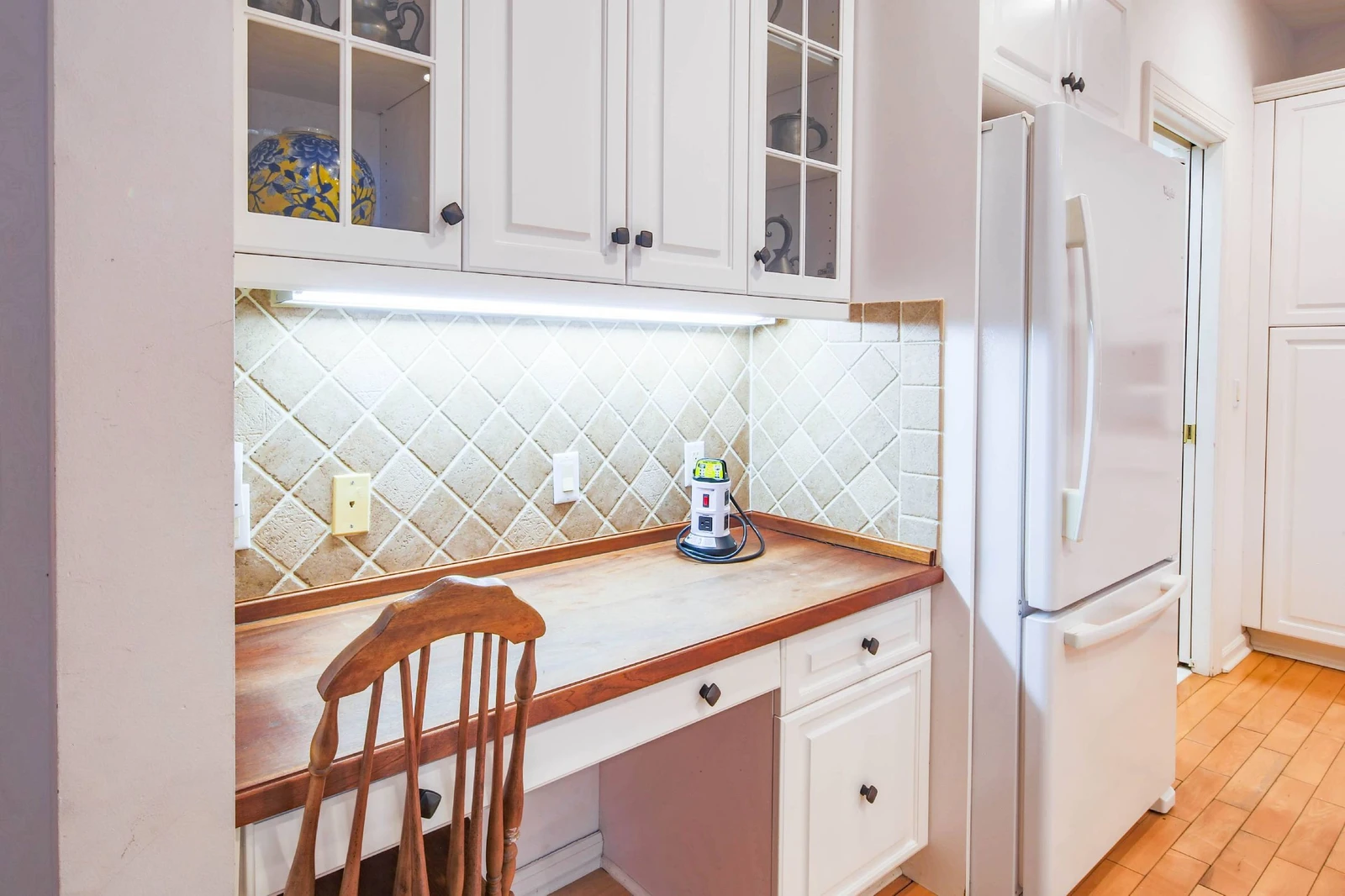 There is a desk in the kitchen near the refrigerator.